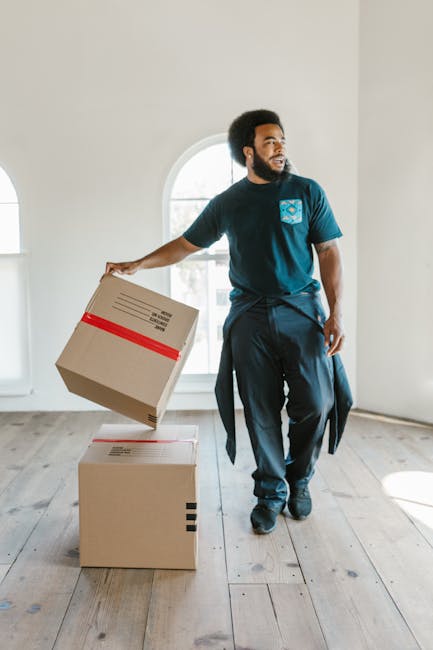 A man with curly black hair and a beard is inside a bright, spacious room with large arched windows allowing natural light to fill the space. He is wearing a teal t-shirt with a logo on the chest and 