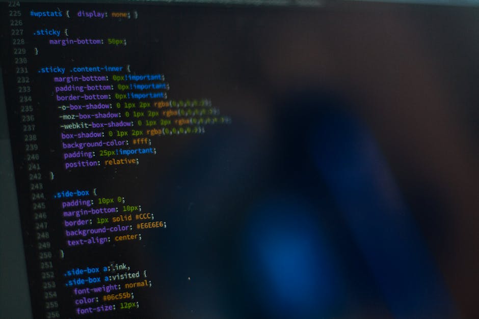 Close-up of a black laptop with a blurred screen displaying lines of code, placed on a dark surface next to a white Puma baseball cap with a black logo. The background is out of focus, emphasizing the